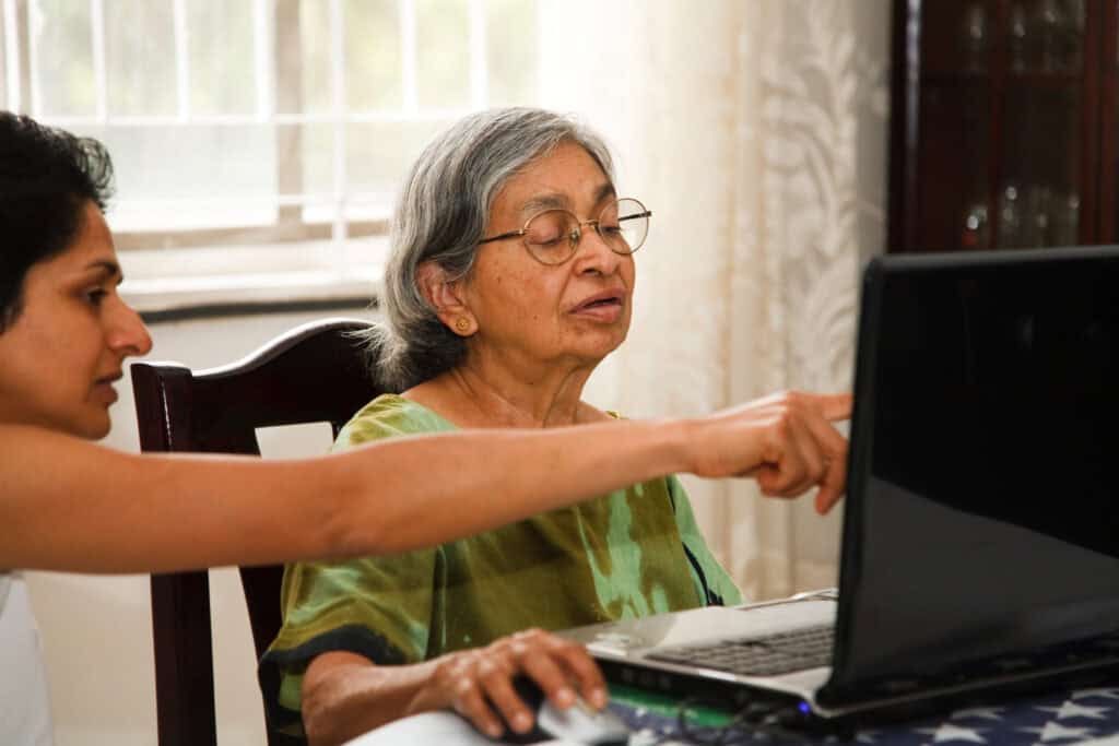 An elderly woman sits at a table using a laptop, while a younger woman beside her points at the screen, assisting her. Sunlight filters through a window in the background.