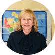 A woman with blonde hair and a dark top is smiling in front of a colorful map of North America on a classroom wall.
