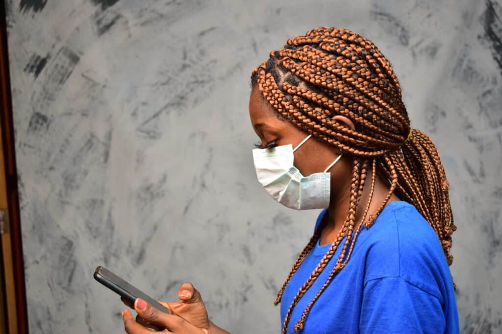 A woman with braided hair wearing a blue shirt and a face mask looks at a smartphone in her hand, standing against a gray, textured background.