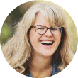 A woman with long, wavy blonde hair and glasses is smiling and laughing. She is wearing a blue shirt and the background is blurred greenery. The photo is cropped in a circular frame.