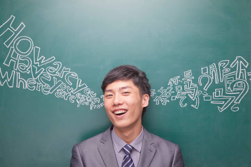 A smiling person in a suit stands in front of a chalkboard with English and Chinese words and phrases like “How are you?” and “Where are you from?” written in chalk around their head.
