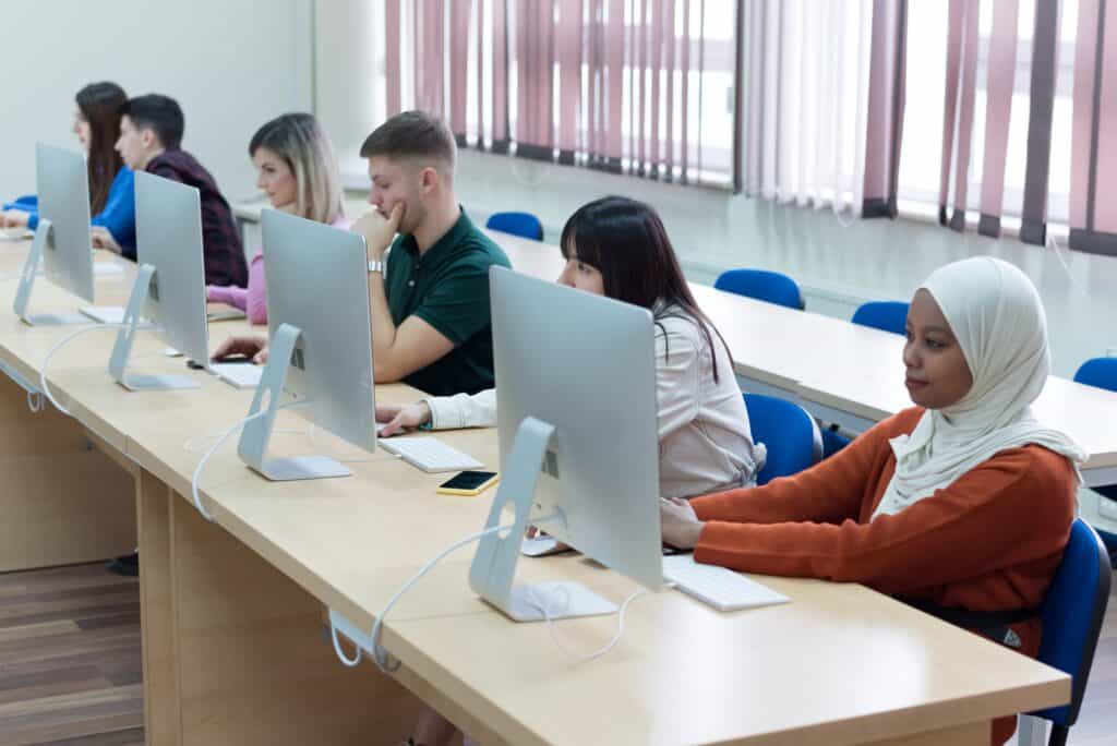 Several students sit in a row at desks using large desktop computers in a classroom. They appear focused on their screens, and the room has large windows with vertical blinds.