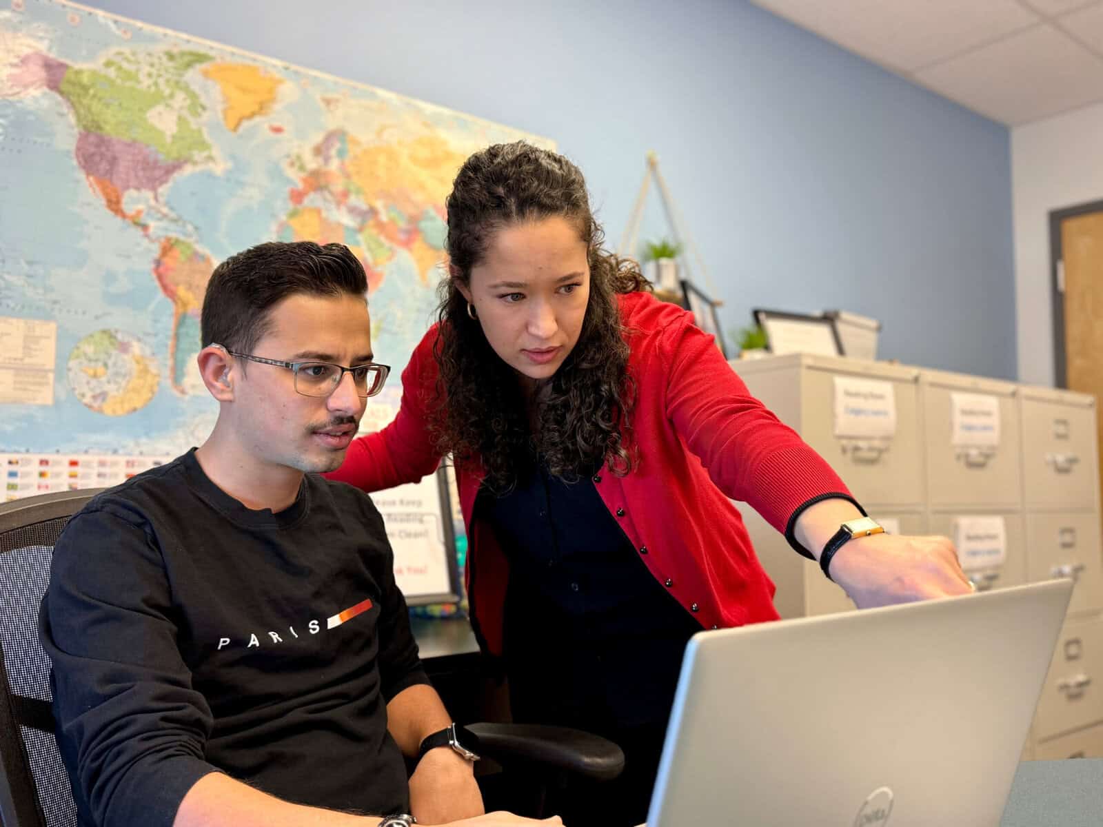 A woman in a red cardigan points at a laptop screen while standing next to a seated man in a black shirt. They appear to be working together at the Literacy Centre of Expertise, with a world map and filing cabinets in the office.