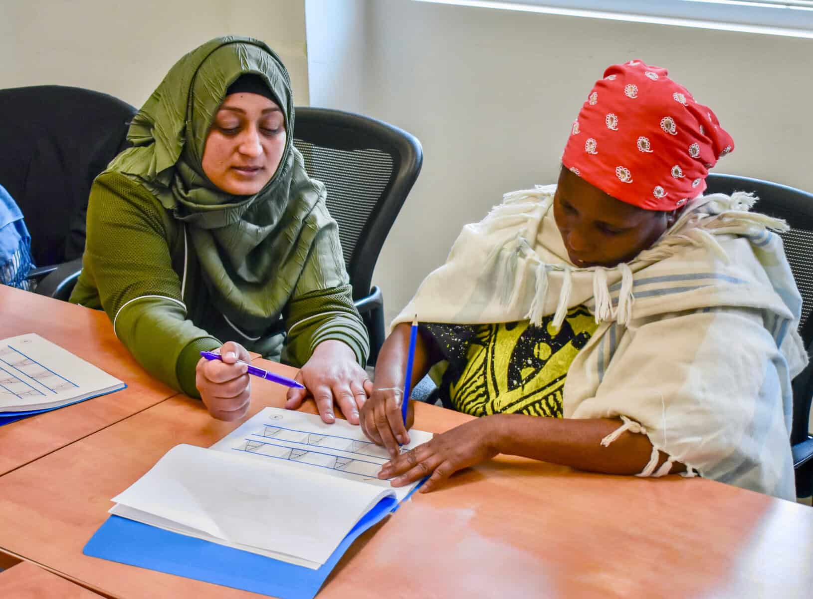 Two women sitting at a table, one in a green hijab and the other in a red headscarf, collaborate on a writing exercise using a worksheet at the Literacy Centre of Expertise. One woman points to the paper while the other writes.