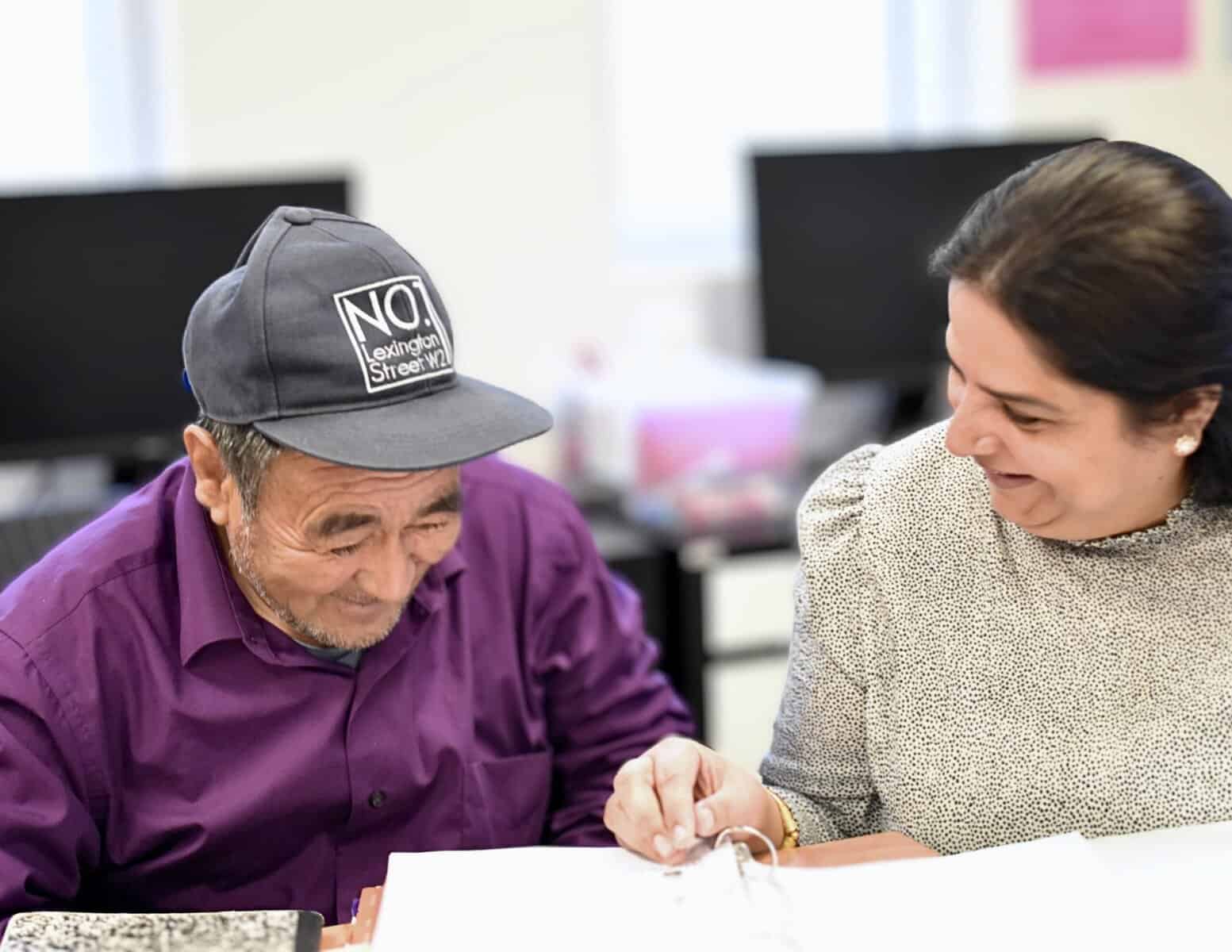 An older man in a purple shirt and cap smiles while looking at papers with a woman in a light patterned blouse, sharing a joyful moment during a discussion at the Literacy Centre of Expertise.