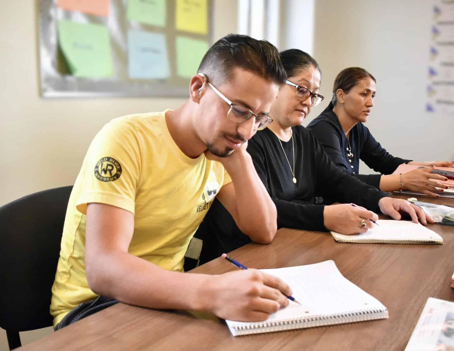 Three adults sit at a table taking notes in notebooks at the Literacy Centre of Expertise. The man in front wears a yellow shirt, and two women sit beside him, all focused on writing. A colorful bulletin board is visible in the background.