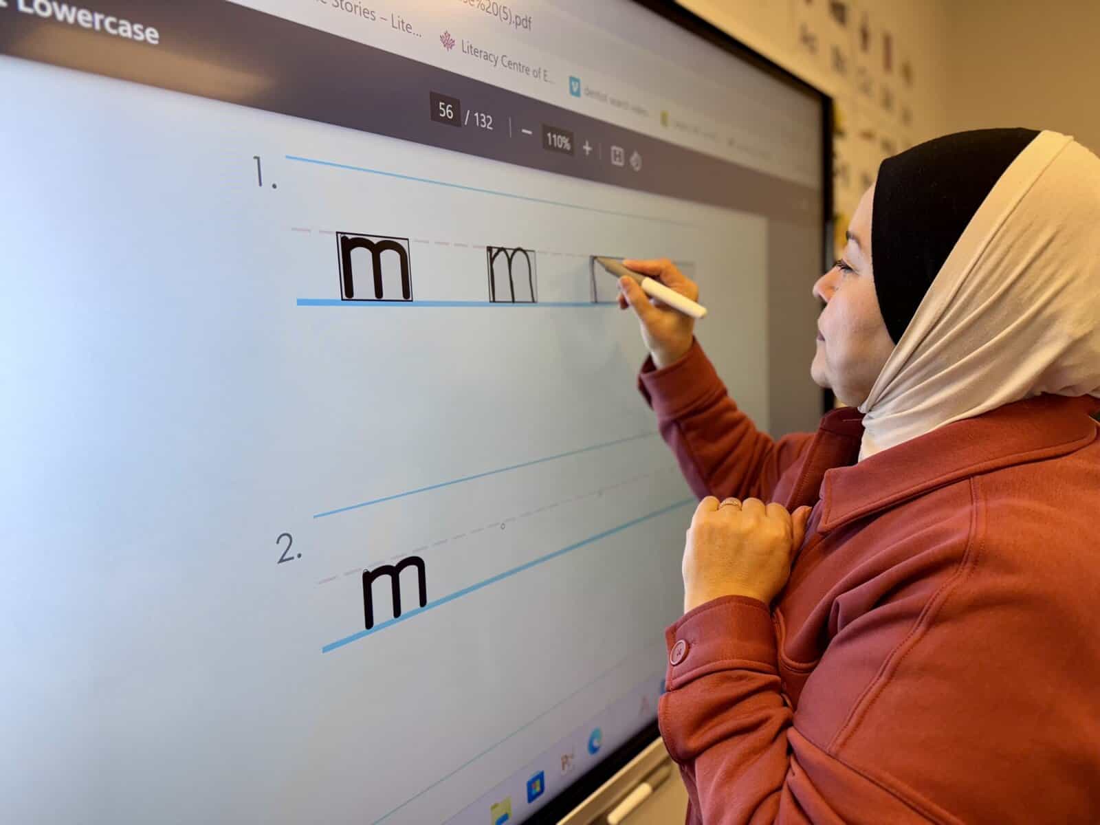 At the Literacy Centre of Expertise, a woman wearing a headscarf practices handwriting with a digital pen, writing the lowercase letter "m" on a large interactive screen.