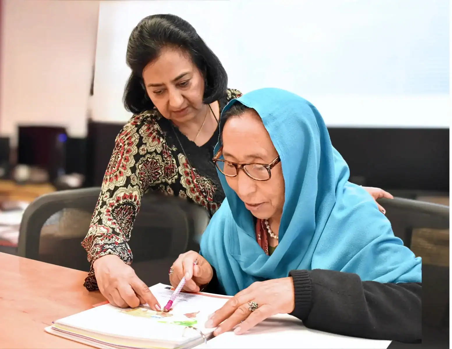Two women sit at a table. One in a blue headscarf points to a book while the other, standing beside her, helps guide and teach—a moment that reflects the supportive spirit of our About Us story.