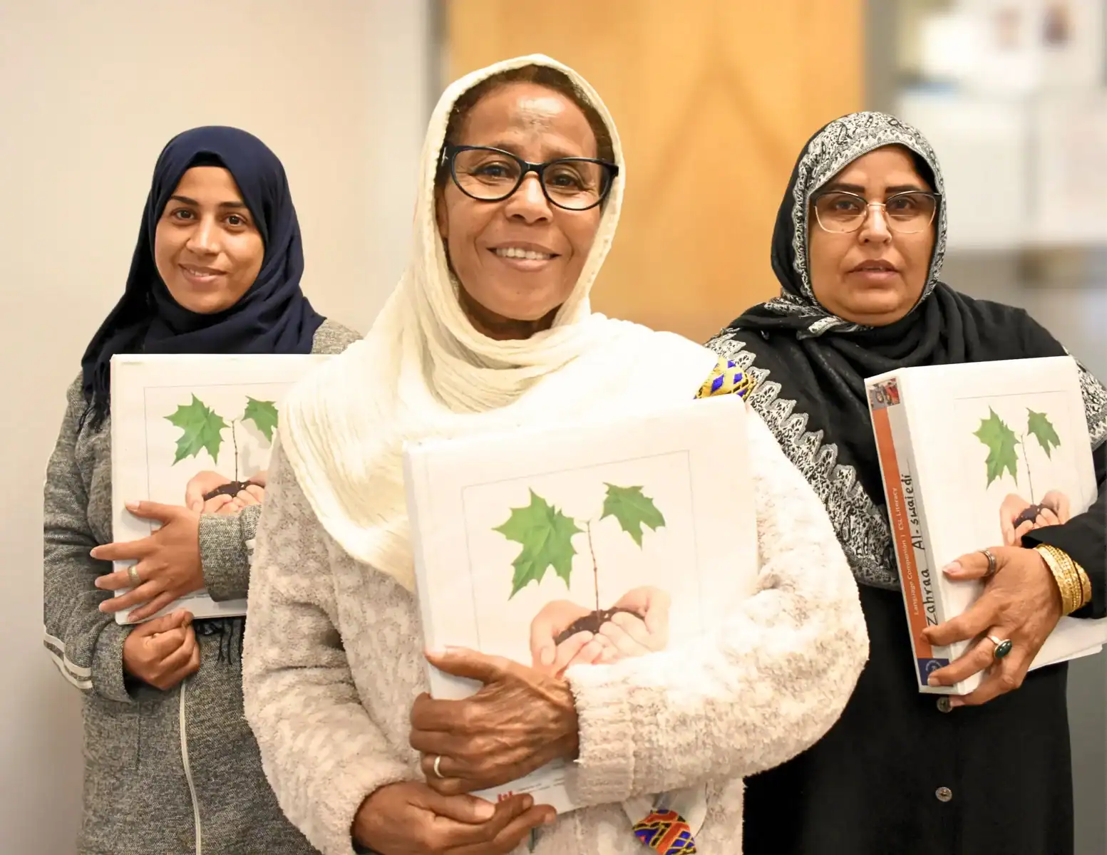 Three women in headscarves smile while holding binders featuring hands nurturing a plant. They stand indoors at the Literacy Centre of Expertise, reflecting an educational or professional environment.