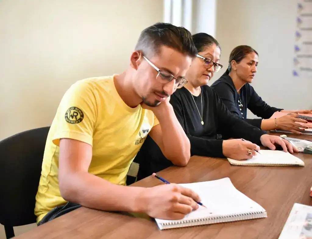 Three adults sit at a table, writing in notebooks. The man in front wears a yellow shirt and glasses, while the two women behind him wear dark clothing. They appear focused and engaged as they use their resources for work.