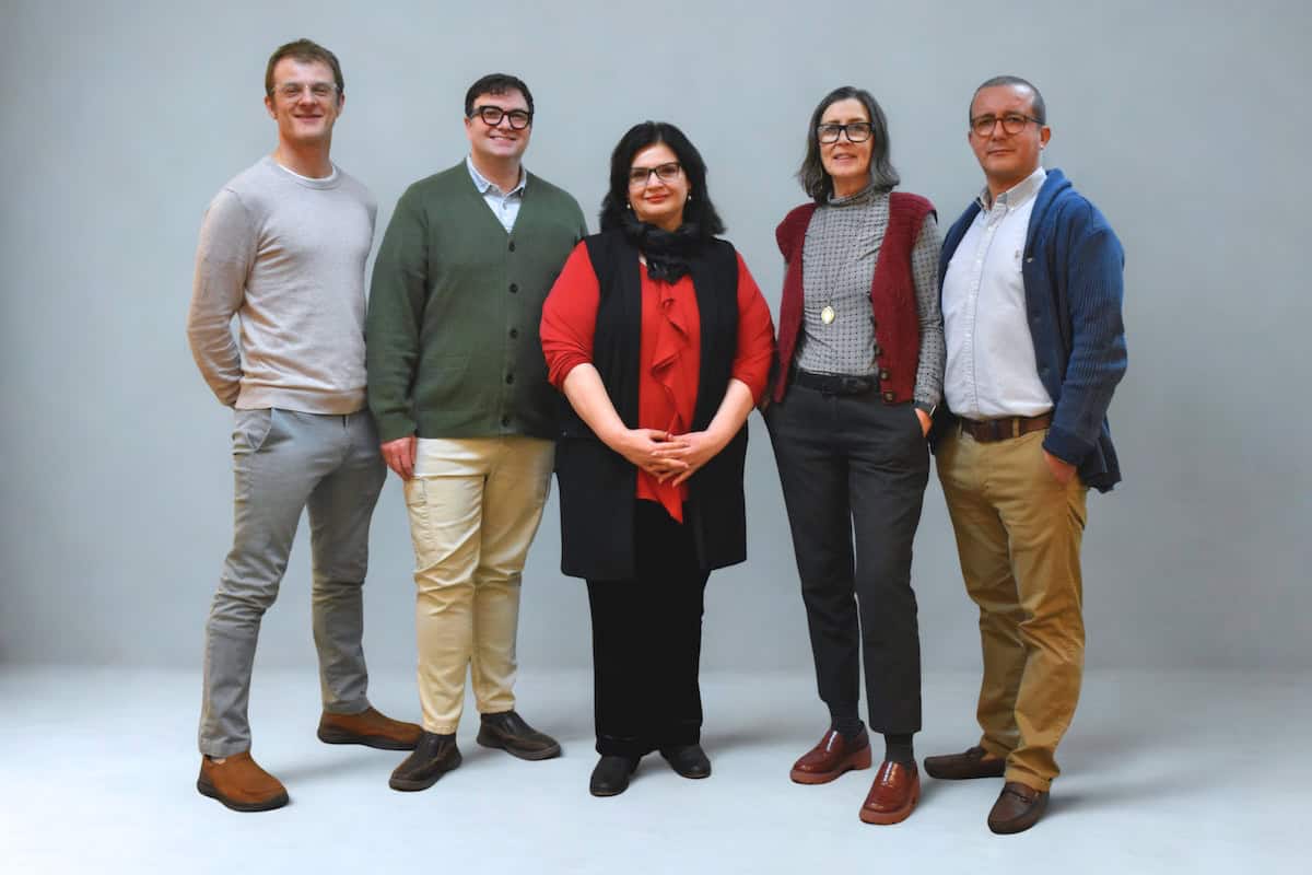 Five adults stand side by side, smiling at the camera against a plain gray background. Dressed in smart-casual clothing, they represent our friendly and professional About Us team in a range of neutral and warm colors.